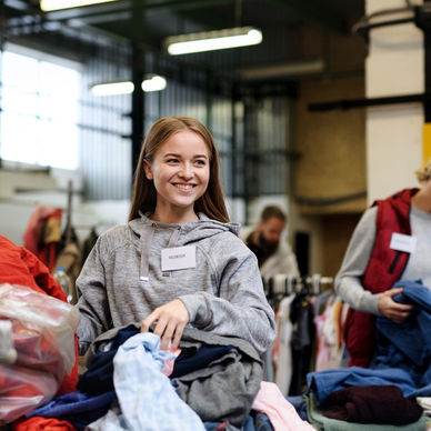 A close-up of a young woman sorting through a stack of clothing with other volunteers at a donation center.