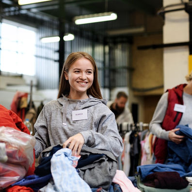 A close-up of a young woman sorting through a stack of clothing with other volunteers at a donation center.
