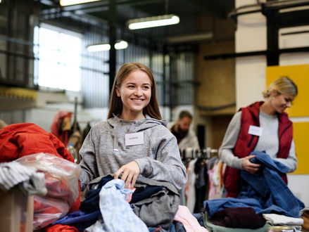 A close-up of a young woman sorting through a stack of clothing with other volunteers at a donation center.