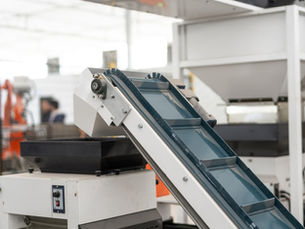 A close-up of the top part of an incline conveyor with a metal belt, next to a large machine inside an industrial room.