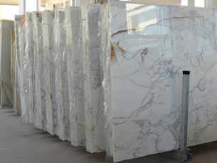 A long row of tall marble slabs standing upright against a metal bar in a large, brightly lit warehouse.