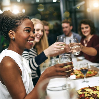 A group of people smiling and raising wine glasses around a table with pizza boxes in a bright indoor room.