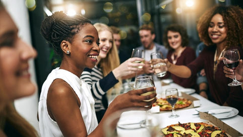 A group of people smiling and raising wine glasses around a table with pizza boxes in a bright indoor room.