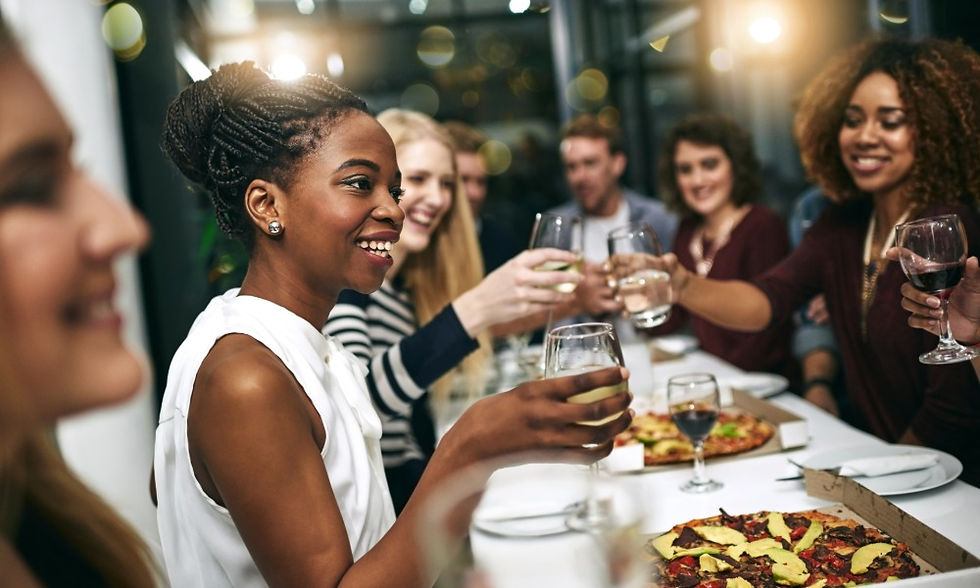A group of people smiling and raising wine glasses around a table with pizza boxes in a bright indoor room.
