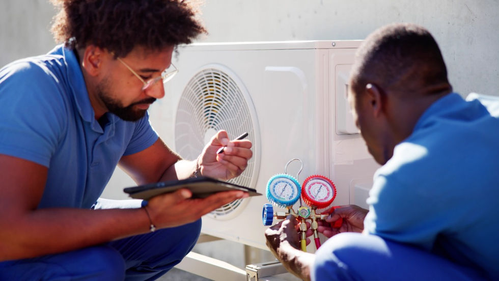 Two men in blue uniforms crouching beside an air conditioning unit, with one holding a tablet and gauges nearby.