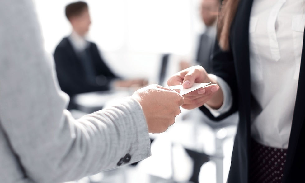 One person hands their business card to another person. Two other people are sitting in chairs nearby.