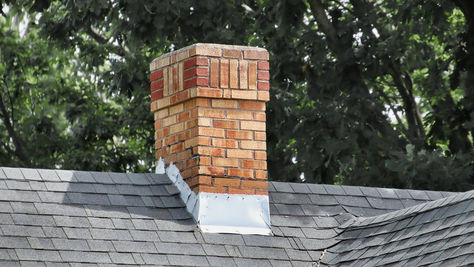 A brick chimney rising from a gray shingled roof. There are trees behind the roof, leaving shade on the chimney.