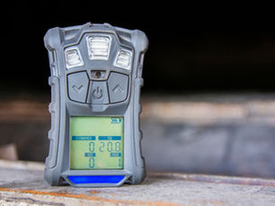 A gray handheld gas detector sits upright on a dirty metal surface. The detector has a digital display.