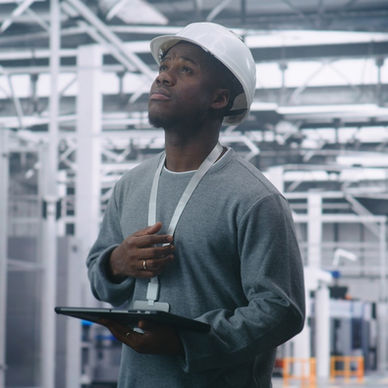 A Black man wearing a white hard hat and lanyard gazes upward while holding a tablet in an industrial facility.