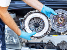 A close-up of a man with blue latex gloves holding up the used pressured plate and clutch disc in front of a car.