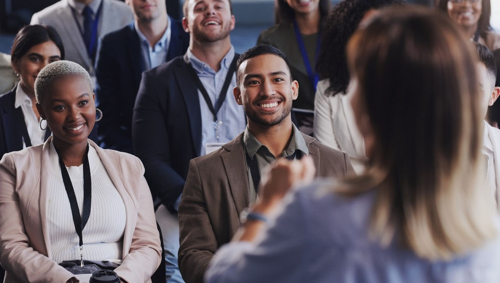 A group of smiling business people all sitting and watching a business speaker that is presenting in front of them.