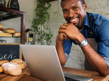 A man smiling while leaning on the front counter of a bakery. There is a laptop and a board with bread in front of him.