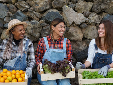 Three smiling women in denim overalls sit in front of a stone wall, each holding a crate filled with produce.