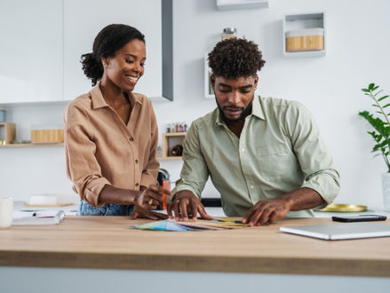 A smiling woman and a man examining colorful paint swatches on a kitchen counter, with a notebook and plant nearby.