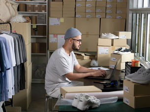 A man sits at a desk in a room filled with cardboard boxes. The man is using a laptop that is sitting on the desk.