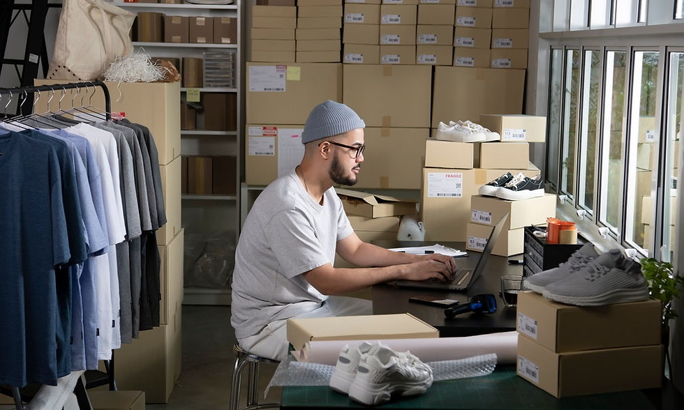 A man sits at a desk in a room filled with cardboard boxes. The man is using a laptop that is sitting on the desk.