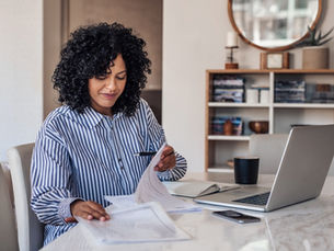 A woman of color with large, curly, black hair, is sitting at her dining room table, going through paperwork.