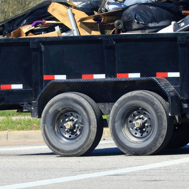 A small open trailer loaded with black garbage bags and loose refuse, parked on an asphalt road outdoors.