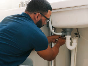 A man wearing glasses and a blue shirt working on white pipes beneath a sink with an open cabinet beside him.
