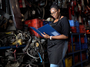 A woman in glasses and a black apron reading a blue folder beside stacked car parts and storage bins.