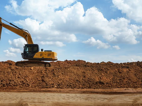 A panoramic view of a yellow excavator on top of dirt and soil underneath a bright, blue, cloudy sky.
