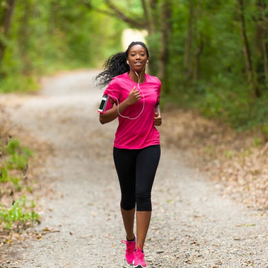 A woman wearing earphones and athletic clothing jogging outdoors on a tree-lined path in bright daylight.