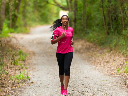 A woman wearing earphones and athletic clothing jogging outdoors on a tree-lined path in bright daylight.