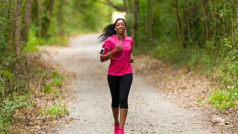 A woman wearing earphones and athletic clothing jogging outdoors on a tree-lined path in bright daylight.