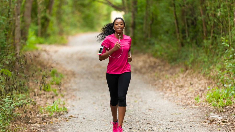 A woman wearing earphones and athletic clothing jogging outdoors on a tree-lined path in bright daylight.