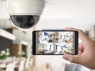 A close-up of a hand holding up a smartphone with views from four home security cameras next to a ceiling-mounted camera.