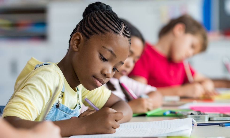 A close-up of a young Black girl writing in a notebook. She's near other students sitting at a table.