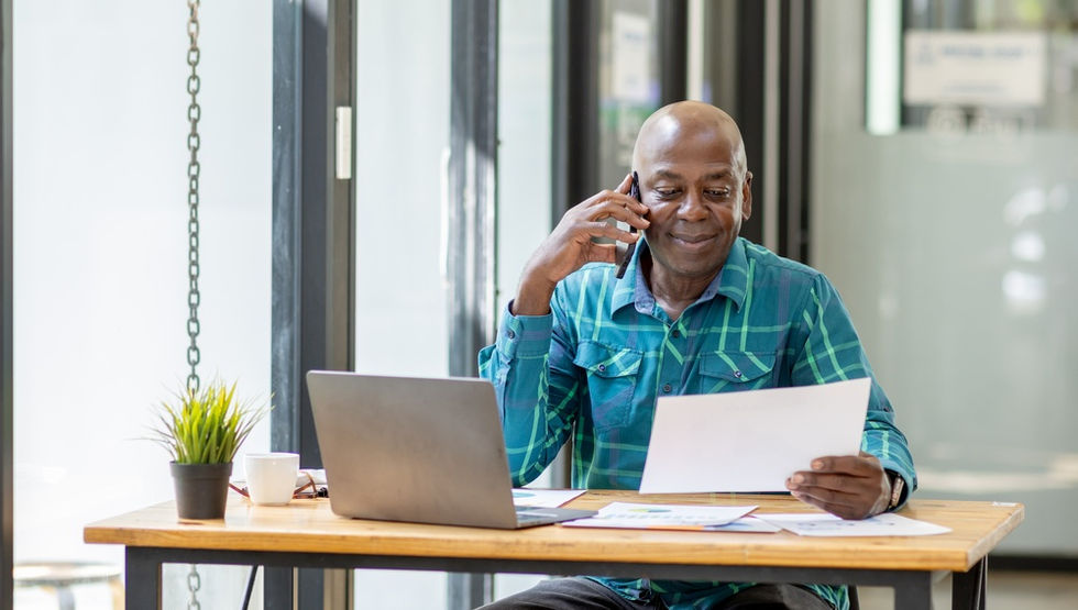 A senior man sitting at a desk and smiling as he looks down at a paper in his hands. He is in the middle of a phone call.