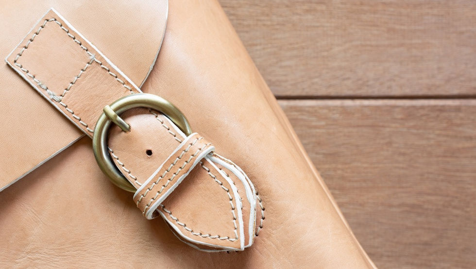 A close-up shows a leather bag strap with a metal buckle and stitching details resting diagonally on a wooden surface.