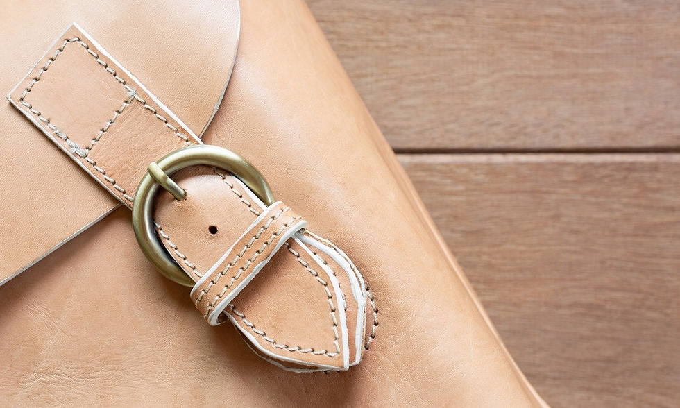 A close-up shows a leather bag strap with a metal buckle and stitching details resting diagonally on a wooden surface.