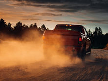 A pickup truck driving along a rural road, kicking up dust at sunset, with grass and trees lining the path.