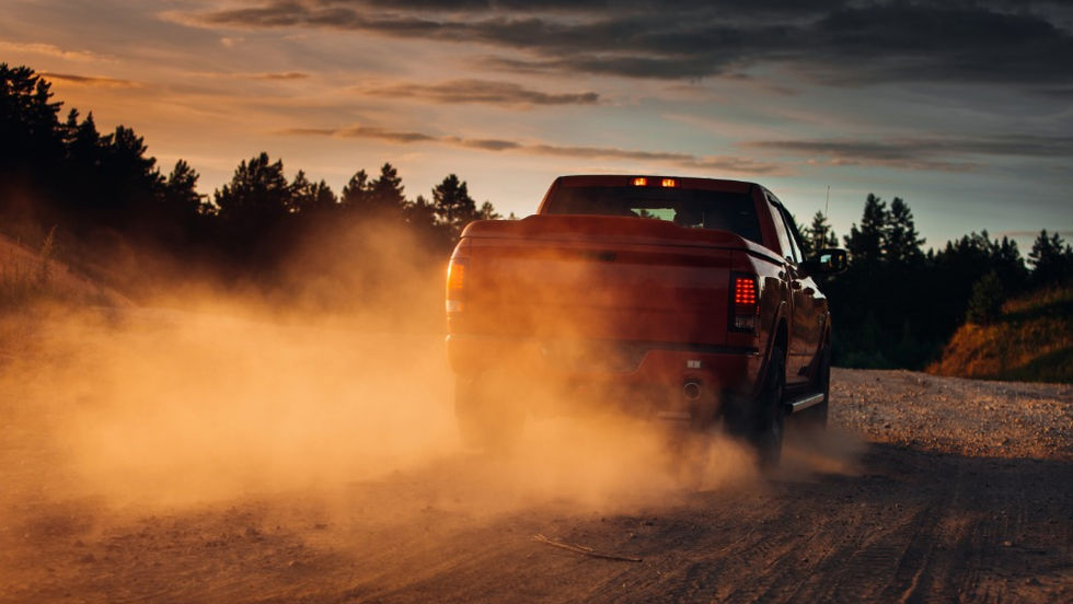 A pickup truck driving along a rural road, kicking up dust at sunset, with grass and trees lining the path.
