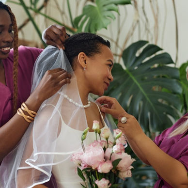 A woman in a white dress holding pink flowers while two women in magenta dresses adjust her veil and necklace.