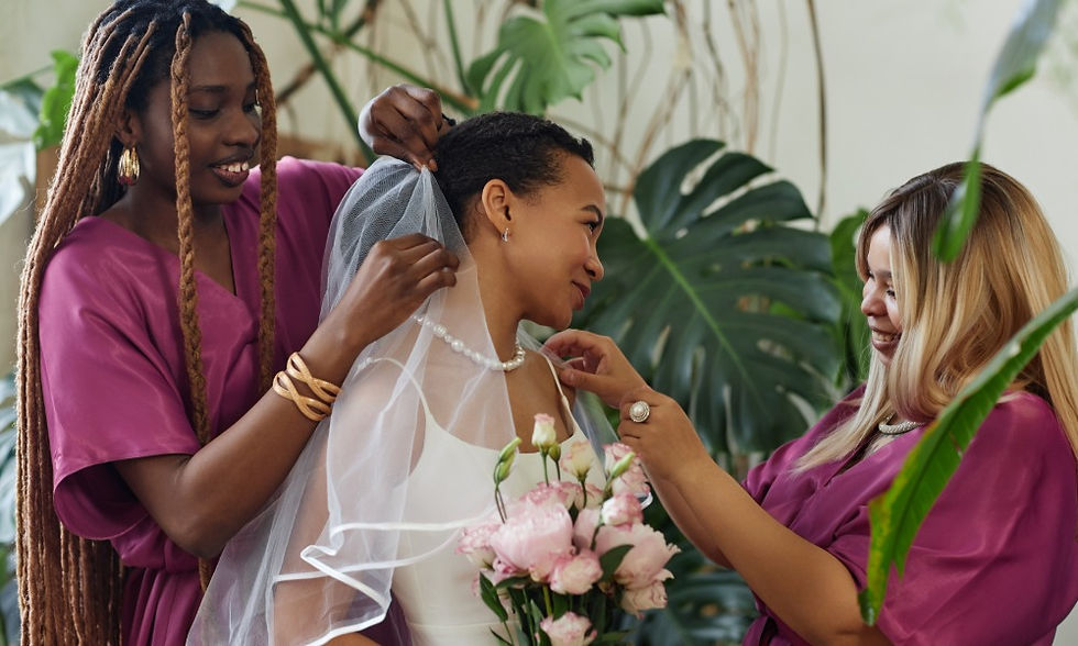 A woman in a white dress holding pink flowers while two women in magenta dresses adjust her veil and necklace.