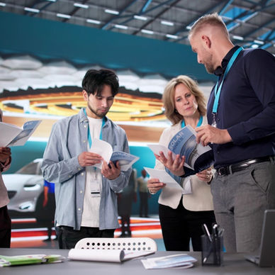 People wearing lanyards are looking at brochures that are part of a trade show booth. The table has a laptop and papers.