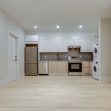 A bright basement apartment contains a modern kitchen, pale wood floors, and a stacked washer and dryer.