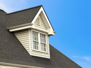 Dormer window on a gray shingled house roof against a bright blue sky.
