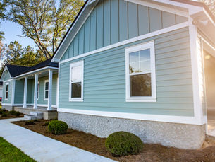 Sunlit blue-gray house exterior with white trim, porch columns, and shrubs along a sidewalk in a quiet suburban yard