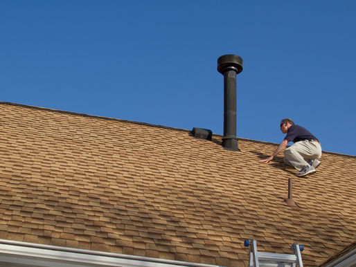 Man on a brown shingle roof, near a chimney, with ladder below. Clear blue sky, suggesting a bright, sunny day.