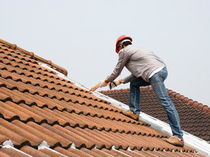 Worker in red helmet applies sealant on brown tiled roof. Clear sky background. Safety gear visible. Focused and careful mood.