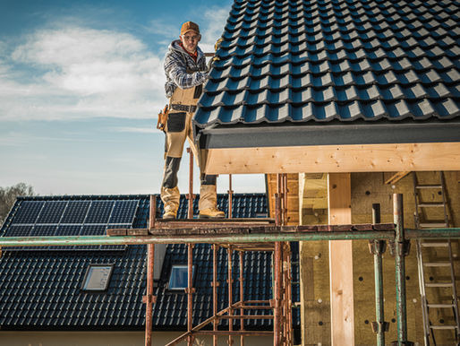 Roofer in overalls roof installation works on a clay tile roof atop scaffolding. Blue sky, solar panels, and another roof are visible in the background. 