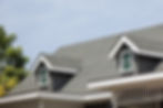 Gray shingled roof with dormer windows on a sunny day; white trim and green trees in the background. Calm and peaceful setting.