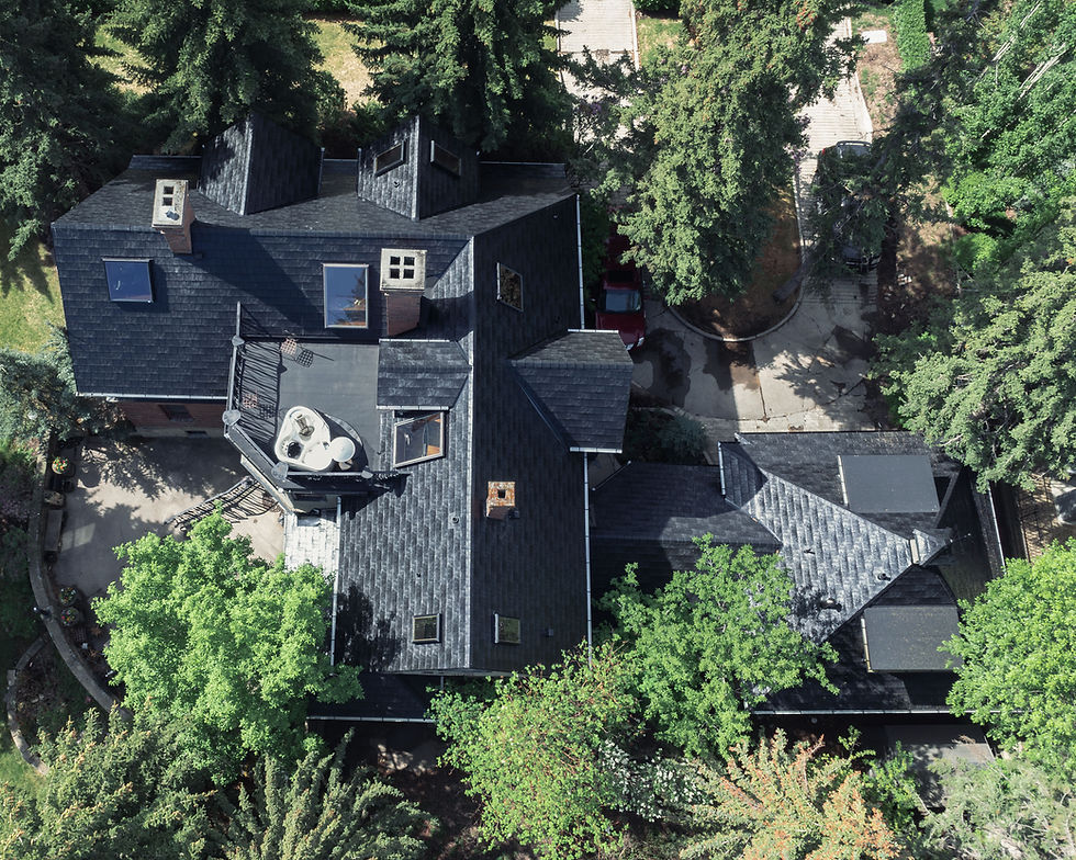 Aerial view of a dark-roofed house nestled among tall trees, with driveways and parked cars, in bright daylight.