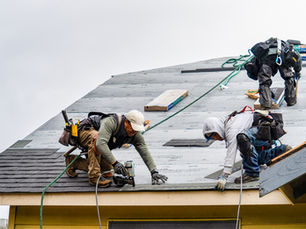 Three workers install roofing shingles on a house. They wear safety gear and use tools. The sky is overcast, creating a focused mood.