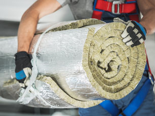 Worker holding a large roll of insulation material. Wearing gloves and a harness, set in an industrial environment. Blue and gray tones.