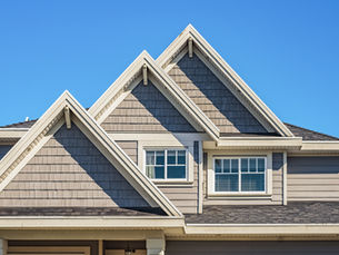 Beige suburban house roof with three steep gables and windows against a clear blue sky.